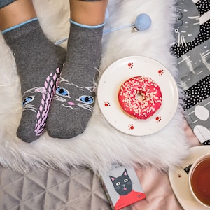 Pode incluir: Um par de meias cinzas com um design de rosto de gato, um donut rosa com granulado branco em um prato branco com pegadas vermelhas, uma xícara de chá e um livro com uma ilustração de gato preto.
