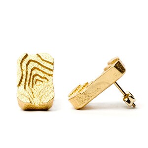 Puede incluir: Un par de pendientes de botón dorados. Cada pendiente tiene forma rectangular con superficie texturizada y diseño de líneas concéntricas. Los pendientes se muestran sobre un fondo blanco.