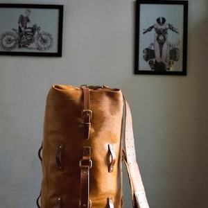 Puede incluir: Una gran bolsa de lona de cuero marrón con múltiples correas y hebillas. La bolsa tiene una forma cilíndrica y está hecha de cuero grueso y duradero.