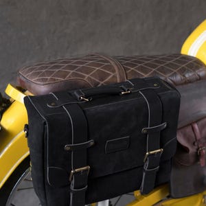 May include: Black leather motorcycle saddlebag with buckles and straps. The bag is attached to a yellow motorcycle.