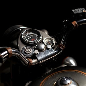 May include: Close-up of a motorcycle's handlebars and instrument panel against a black background. The speedometer reads 26018. The handlebars are wrapped with brown leather straps. The motorcycle has a silver finish with orange accents.