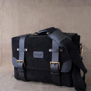 May include: Black leather messenger bag with a suede finish, featuring two adjustable straps with brass buckles and a top handle. The bag has a front flap with a brass closure and a brand label that reads "remarkable".