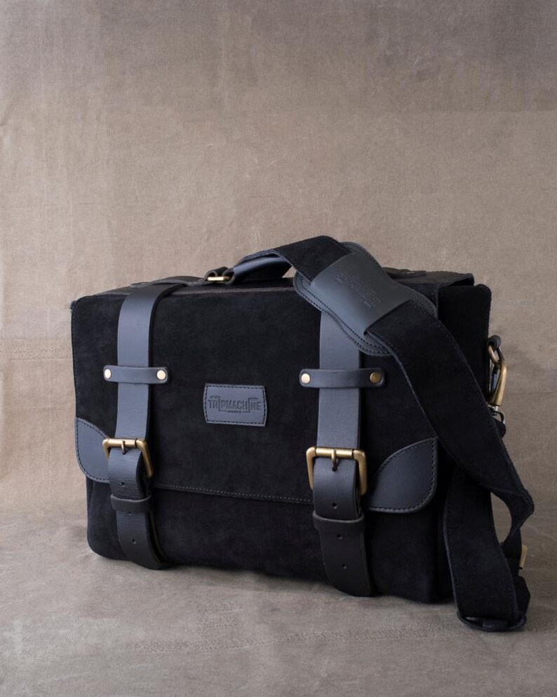 May include: Black leather messenger bag with a suede finish, featuring two adjustable straps with brass buckles and a top handle. The bag has a front flap with a brass closure and a brand label that reads "remarkable".