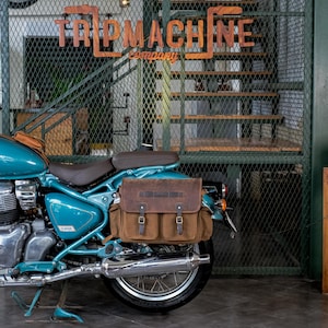 May include: A teal motorcycle with a brown leather saddlebag is parked in front of a metal gate. The saddlebag has two front pockets and is secured with buckles. The sign above the gate reads "TRIP MACHINE company".