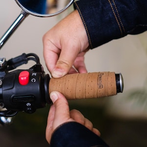 May include: A brown leather motorcycle handlebar grip with the text "Tripmachine" embossed on it. The grip is being installed on a motorcycle handlebar.