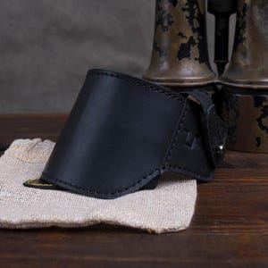 May include: A black leather arm guard with a curved design, featuring a stitched edge and an elastic strap. It rests on a beige fabric pouch with a yellow trim. The background includes antique brass binoculars.