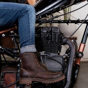 May include: Brown leather work boots with a lug sole and yellow stitching. The boots are worn by a person sitting on a black motorcycle. The motorcycle has copper-colored accents and the words "The Anchor" with an anchor logo.