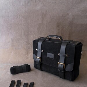 May include: A black leather briefcase with a top handle and multiple buckles. The bag has a small leather patch with the word "Genuine" on it. The bag is accompanied by a black strap and three black straps with metal clips.