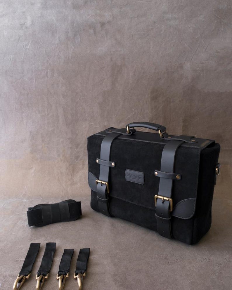 May include: A black leather briefcase with a top handle and multiple buckles. The bag has a small leather patch with the word "Genuine" on it. The bag is accompanied by a black strap and three black straps with metal clips.