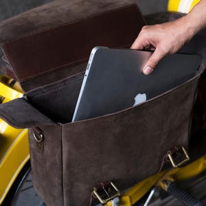 May include: A brown leather motorcycle saddlebag with a laptop computer inside. The bag has a flap with a buckle closure and a strap with a buckle. The bag is attached to a yellow motorcycle.