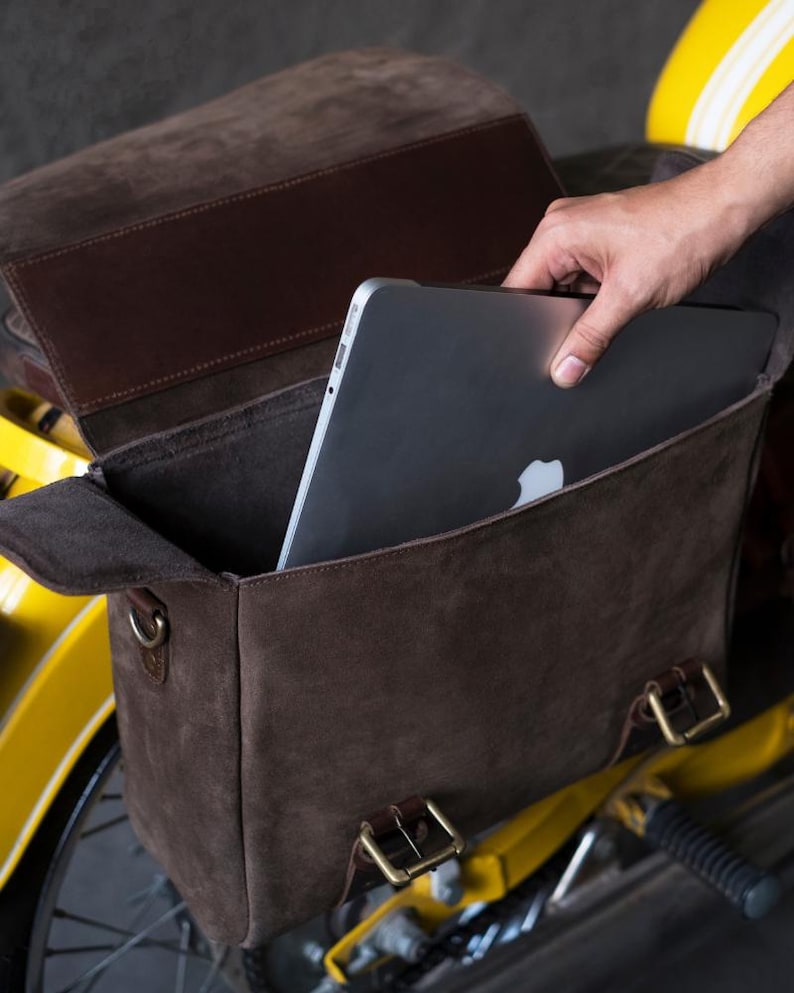 May include: A brown leather motorcycle saddlebag with a laptop computer inside. The bag has a flap with a buckle closure and a strap with a buckle. The bag is attached to a yellow motorcycle.