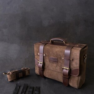 May include: A brown leather briefcase with a top handle, two buckles, and a detachable shoulder strap. The bag has a brown suede exterior and a dark brown leather trim. The bag is sitting on a gray surface with four black straps with brass hooks.