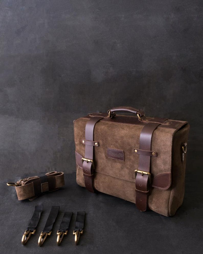 May include: A brown leather briefcase with a top handle, two buckles, and a detachable shoulder strap. The bag has a brown suede exterior and a dark brown leather trim. The bag is sitting on a gray surface with four black straps with brass hooks.