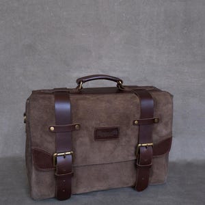 May include: A brown leather briefcase with two buckles and a top handle. The bag has a brown suede exterior and a brass nameplate that reads "Remarkable".
