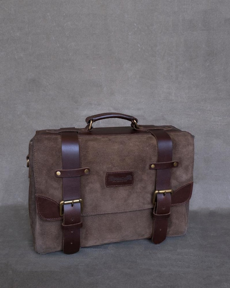 May include: A brown leather briefcase with two buckles and a top handle. The bag has a brown suede exterior and a brass nameplate that reads "Remarkable".