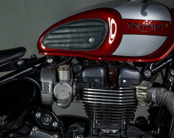 May include: Close-up of a Triumph motorcycle engine and fuel tank. The tank is red and silver with a black leather seat. The engine is silver with black accents. The word "Triumph" is visible on the tank.