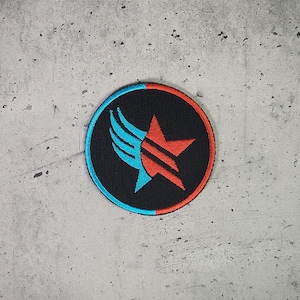 Può includere: Un cerotto circolare bianco e nero con una stella rossa e ali blu e arancioni. Il cerotto è su uno sfondo grigio.