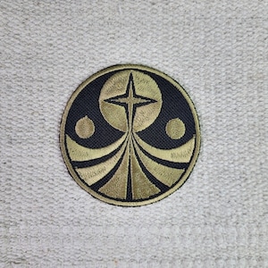 Può includere: Una toppa ricamata circolare con sfondo nero e cuciture dorate. Il design presenta una croce stilizzata con due cerchi su entrambi i lati, il tutto all'interno di un cerchio.