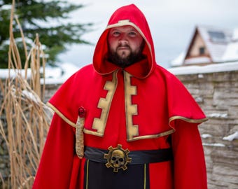 Può includere: Un mantello rosso con cappuccio con finiture dorate e una cintura nera con fibbia a teschio. Il costume ha un tema medievale o fantasy. La persona indossa un cappuccio e un mantello rossi.