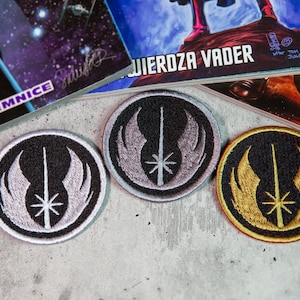 Puede incluir: Tres parches bordados con el símbolo de la Alianza Rebelde de Star Wars. Los parches son circulares y tienen un fondo negro con bordados blancos, plateados y dorados. El símbolo presenta un sable de luz, alas y una estrella.
