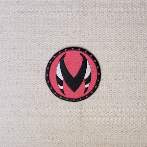 May include: A red and black embroidered patch featuring a stylised V shape in the centre. The patch is circular and has a black border with small white dots.