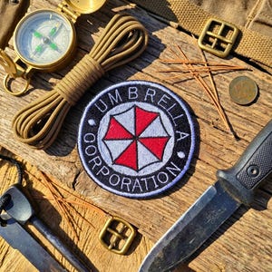 Może przedstawiać: Asortyment sprzętu survivalowego, w tym kompas, zwinięta beżowa lina, czarny nóż i naszywka z napisem "UMBRELLA CORPORATION" oraz logo czerwono-białego parasola. Przedmioty ułożone na zwietrzałej drewnianej powierzchni.