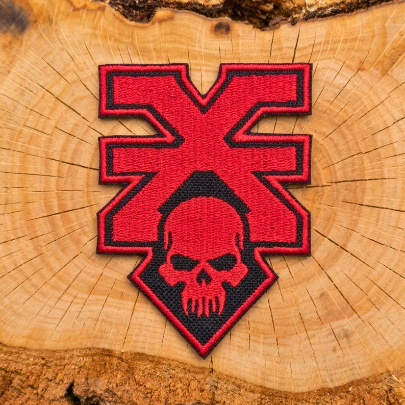 May include: A red and black embroidered patch with a skull design. The patch has a geometric shape with a skull and crossbones design. The patch is on a wooden surface.