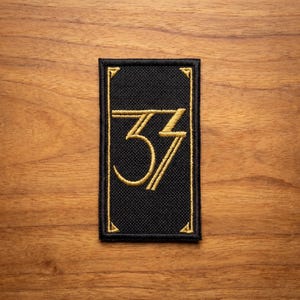 May include: Black rectangular patch with gold embroidered number "33" in a stylized art deco font. The patch has a gold border and is set against a wooden background. The patch is rectangular.