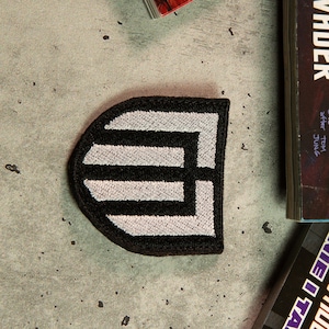 May include: Black and white embroidered patch in the shape of a shield with a geometric design.