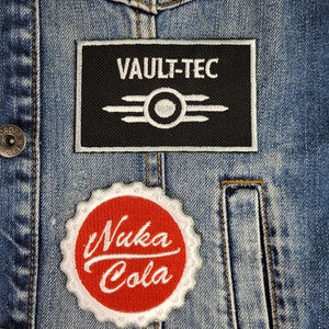 Peut inclure: Une veste en jean avec deux patchs. Le patch du haut est noir avec du texte blanc qui dit "VAULT-TEC" et un logo Vault-Tec blanc. Le patch du bas est blanc avec un cercle rouge qui dit "Nuka Cola" en blanc.