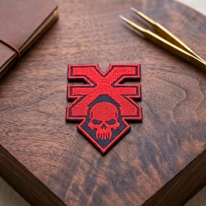 May include: A red and black embroidered patch with a skull design and the letter "X". The patch is on a dark wooden surface, with a leather notebook and tweezers in the background. The patch has a detailed design.