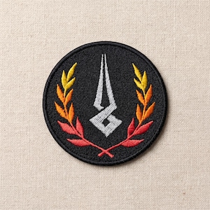 Könnte beinhalten: Runder, schwarzer, gestickter Aufnäher mit einem weißen, stilisierten Emblem. Das Emblem wird von einem Lorbeerkranz in Gelb-, Orange- und Rottönen flankiert. Geeignet für Kleidung oder Accessoires.