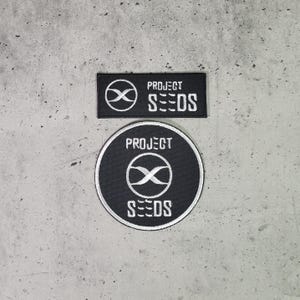 Op de afbeelding: Twee geborduurde patches in zwart en wit met de tekst "PROJECT SEEDS" en een gestileerd oneindigheidssymbool.