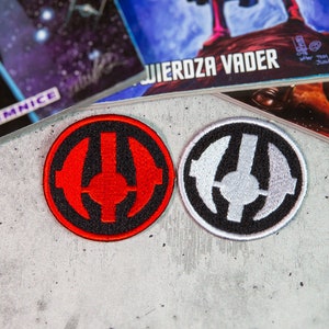 Może przedstawiać: Dwa haftowane naszywki, jedna czerwona, a druga biała, z czarnym tłem i stylizowanym symbolem. Symbol to koło z pionową linią w środku i dwoma zakrzywionymi liniami po obu stronach.