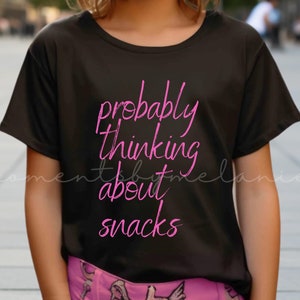 Pode incluir: Uma pessoa jovem vestindo uma camiseta preta com o texto "probably thinking about snacks" em letras cor-de-rosa.