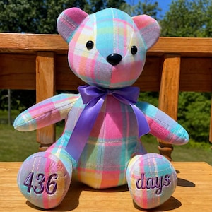 May include: A plush teddy bear made of pastel plaid fabric in shades of pink, blue, yellow, and white. The bear features a purple satin ribbon tied around its neck, black button eyes, and a black nose. The number 436 is embroidered on the left foot.