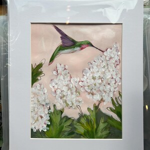 Puede incluir: Pintura enmarcada de un colibrí cerca de flores blancas. El colibrí tiene colores verdes, morados y rojos. Las flores son blancas con hojas verdes, sobre un fondo rosa.