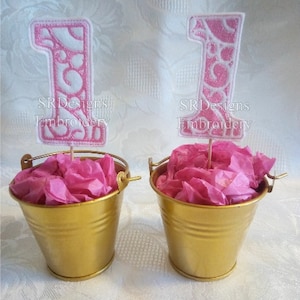 May include: Two gold metal buckets filled with pink tissue paper, each holding a pink and white number one decoration. The number one decorations have a decorative design and the text "SR Designs Embroidery".