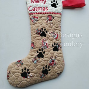 May include: A quilted Christmas stocking with the words "Merry Catmas" embroidered in red. The stocking features black paw prints and a pattern of cats in festive outfits. A red ribbon loop is attached.