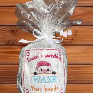 May include: A wrapped gift with a white embroidered design. The design features the text "Santa's watching, Wash Your hands" and a cartoon Santa Claus in a red hat. The gift is wrapped in clear cellophane and tied with a white ribbon.