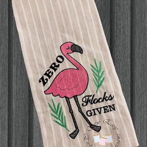Puede incluir: Un paño de cocina a rayas beige y blanco con un diseño de flamenco rosa. El flamenco tiene patas y pico negros, con el texto "ZERO Flocks GIVEN" en negro. También hay hojas verdes y carretes de hilo bordados en el paño.