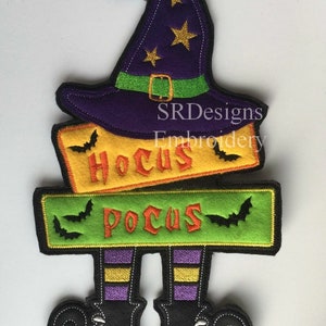 May include: A Halloween-themed felt decoration featuring a purple witch hat with gold stars, a green band, and the words "Hocus Pocus" and "Pocus" on yellow and green banners. Black bat silhouettes and witch legs complete the design.