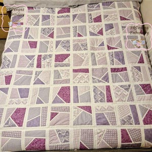 May include: A patchwork quilt with squares and triangles in shades of purple and white. The quilt is made from fabric with different patterns, including floral and geometric designs. The quilt is labelled "SR Designs Embroidery".