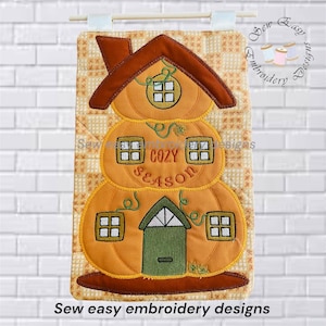 May include: A quilted wall hanging featuring a three-tiered pumpkin house design in shades of orange, brown, and green. The embroidery includes windows, a green door, and the text "COZY SEASON" stitched in the center on a tan patterned background.