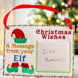Peut inclure: Panneau de Noël en feutre avec un ruban rouge. Il affiche les mots brodés "Christmas Wishes" et "A Message from your Elf". Il comprend un chapeau d'elfe, des chaussures et un bloc-notes avec "I'M BACK!!!".