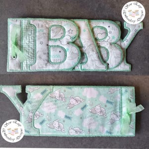 May include: A fabric sign spelling "BABY" in large, padded letters. The letters are white with a pale green trim and feature a pattern of stars, moons, and sleeping baby elephants. A matching fabric piece is below, with a green ribbon.