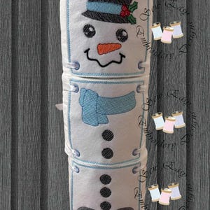 Op de afbeelding: Een wit decoratief item in de vorm van een sneeuwman met een zwarte hoed, blauwe sjaal en wortelneus. De sneeuwman heeft zwarte ogen, een lachende mond en zwarte knopen. Het item is gemaakt van vilt.