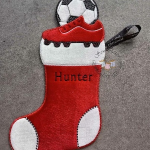 Op de afbeelding: Rood en wit kerstsokornament met een voetbal en een rode voetbalschoen. De naam "Hunter" is op de sok geborduurd. Een zwart lint is bevestigd om op te hangen.