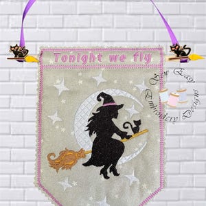 May include: A light grey fabric wall hanging featuring an embroidered silhouette of a witch riding a broomstick in front of a crescent moon. The text Tonight we fly is stitched in pink above the moon. It has a purple ribbon hanger and black cat accents.