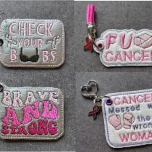 Może przedstawiać: Cztery haftowane breloczki z różowym i białym tekstem i wzorami. Breloczki mają frazy takie jak "CHECK YOUR BBS" i "FU CANCER". Każdy breloczek ma różową frędzlę i czerwoną wstążkę.